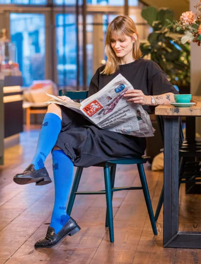 Vrouw leest krant aan een tafel in een restaurant. Ze draagt de blauwe supportkousen van Herzog.